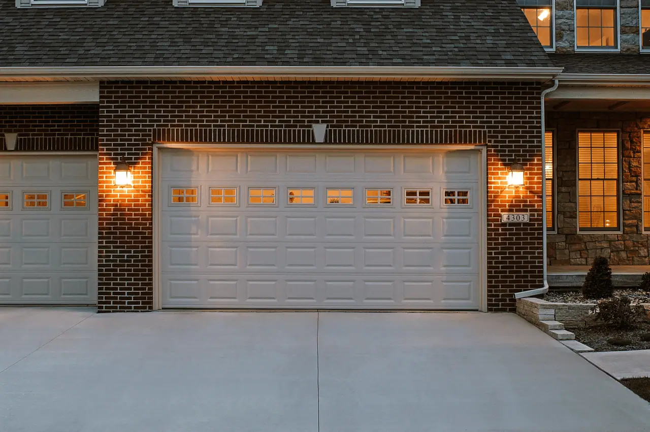 Brick home with a white raised-panel garage door lit by warm wall lanterns at dusk